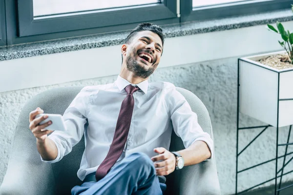 Fröhlicher Geschäftsmann sitzt im Sessel und lacht beim Videochat auf dem Smartphone — Stockfoto