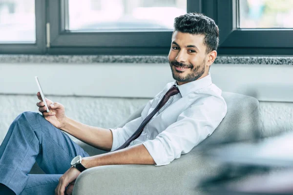 Selektiver Fokus eines gut aussehenden Geschäftsmannes, der in die Kamera lächelt, während er im Sessel sitzt und das Smartphone in der Hand hält — Stockfoto