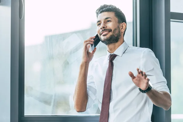 Sonriente hombre de negocios mostrando gesto de espera mientras habla en el teléfono inteligente - foto de stock