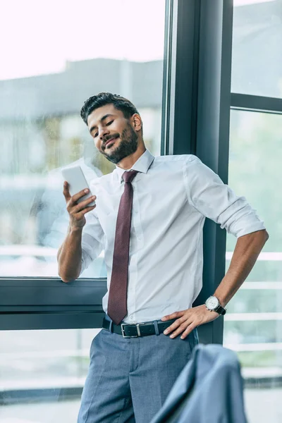 Handsome businessman smiling and holding hand on hip during video chat on smartphone — Stock Photo