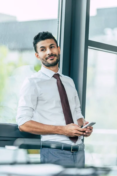 Selective focus of cheerful businessman standing near window, holding smartphone and looking away — Stock Photo