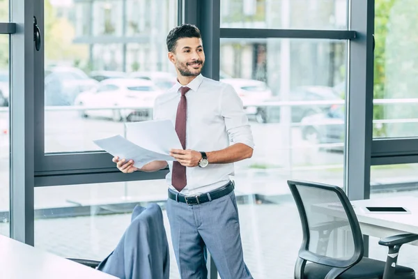 Hombre de negocios guapo sosteniendo documentos, sonriendo y mirando hacia otro lado - foto de stock