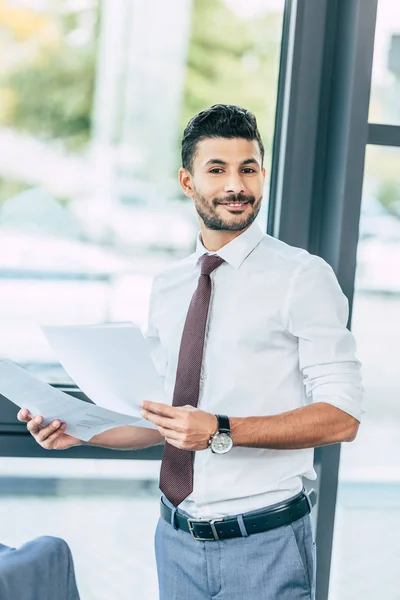 Hombre de negocios guapo sosteniendo documentos, sonriendo y mirando a la cámara - foto de stock