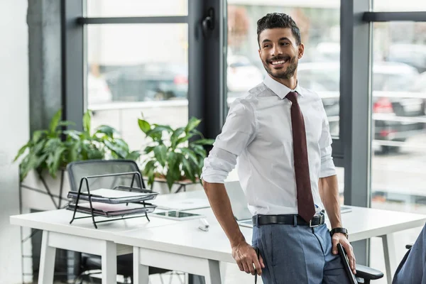 Giovane uomo d'affari sorridente e guardando lontano mentre in piedi sul posto di lavoro — Foto stock