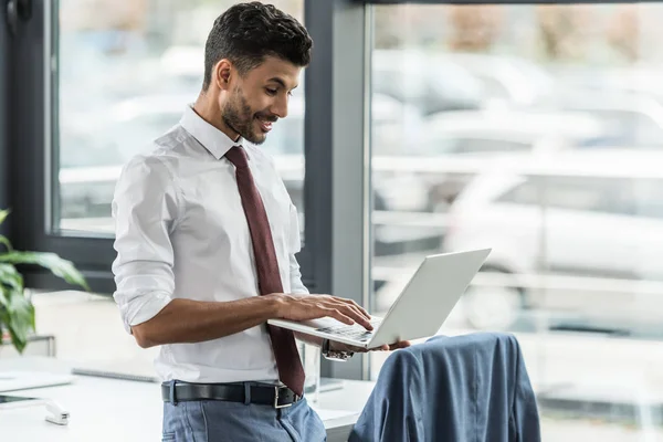 Улыбающийся бизнесмен с помощью ноутбука, стоя на рабочем месте — стоковое фото