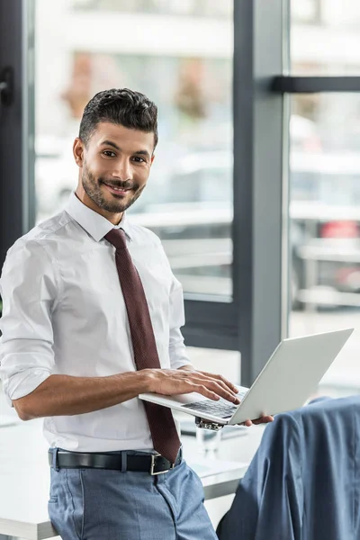 Jovem empresário segurando laptop e sorrindo para a câmera — Fotografia de Stock