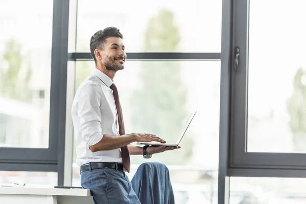 Cheerful businessman holding laptop and looking away in office — Stock Photo