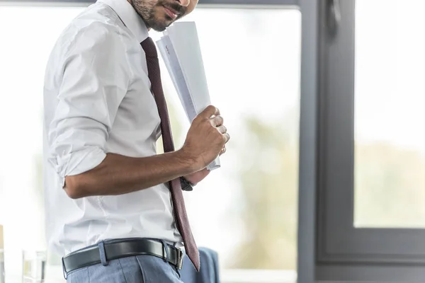 Vista recortada de un hombre de negocios sonriente sosteniendo papeles mientras está parado en el lugar de trabajo - foto de stock