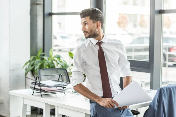 Thoughtful businessman holding papers and looking away while standing at workplace — Stock Photo