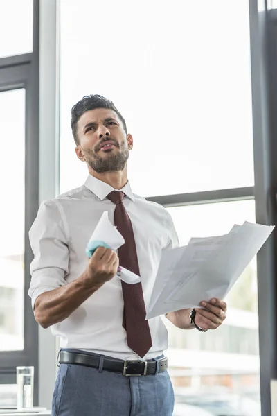 Hombre de negocios insatisfecho arrugando documento mientras está de pie cerca de la ventana en la oficina - foto de stock