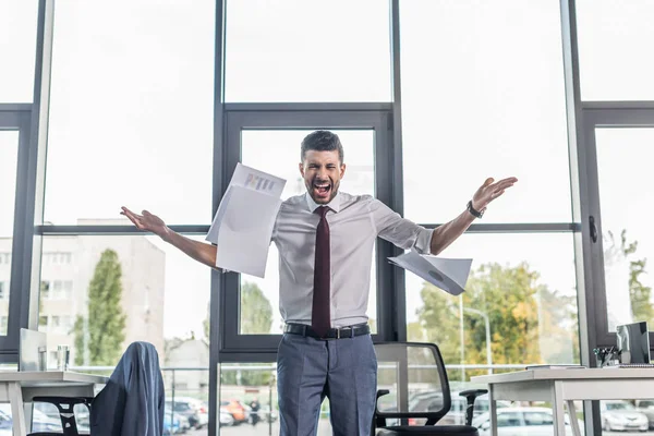 Homem de negócios irritado jogando papéis e gritando enquanto estava no escritório moderno — Fotografia de Stock