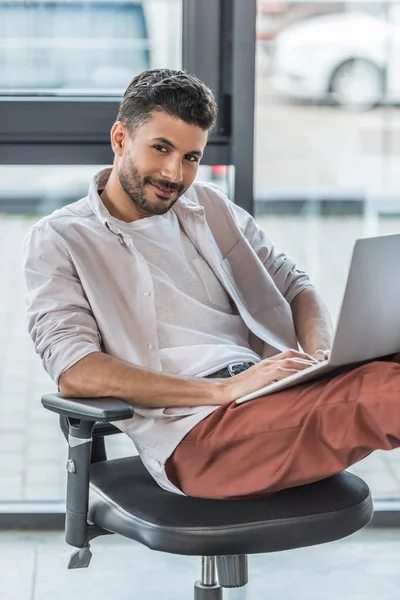 Hombre de negocios sonriente con ropa casual sentado en la silla de la oficina, utilizando el ordenador portátil y mirando a la cámara - foto de stock