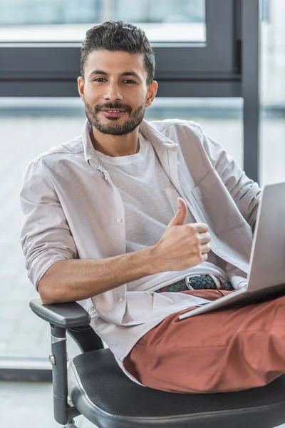 Hombre de negocios sonriente con ropa casual sentado en la silla de la oficina, mostrando el pulgar hacia arriba y mirando a la cámara - foto de stock