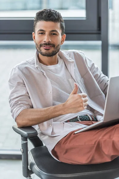 Homme d'affaires souriant en vêtements décontractés assis sur une chaise de bureau, montrant pouce levé et regardant la caméra — Photo de stock