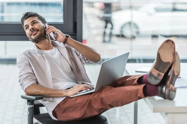 Alegre hombre de negocios sentado con las piernas en el escritorio y hablando en el teléfono inteligente en la oficina - foto de stock