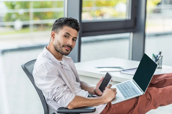 Joven, empresario confiado sosteniendo el ordenador portátil y el teléfono inteligente con pantalla en blanco mientras mira a la cámara - foto de stock