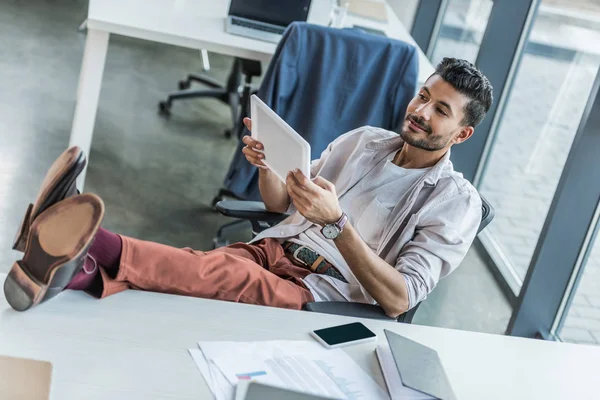 Young businessman using digital tablet while sitting at workplace with legs on desk — Stock Photo