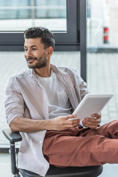 Hombre de negocios sonriente sentado en la silla de oficina, sosteniendo la tableta digital y mirando hacia otro lado - foto de stock