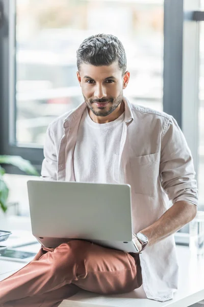 Joven hombre de negocios sonriendo a la cámara mientras está sentado en el escritorio y utilizando el ordenador portátil - foto de stock