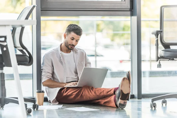 Hombre de negocios concentrado en ropa casual sentado en el suelo cerca de la ventana y utilizando el ordenador portátil - foto de stock
