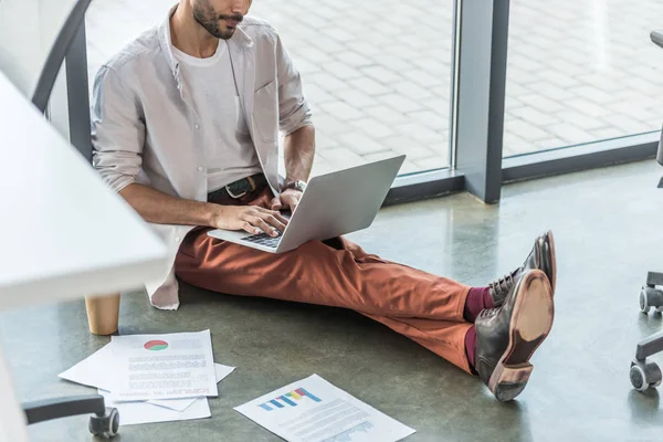 Cropped view of businessman in casual clothes sitting on floor near documents and using laptop — Stock Photo