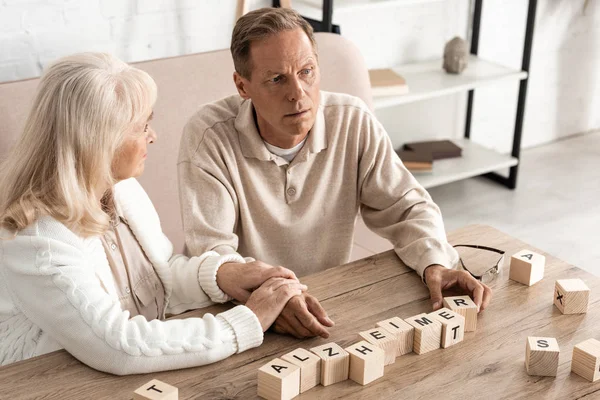 Molesto anciano mujer sentado cerca enfermo marido cerca de madera cubos con alzheimer cartas - foto de stock