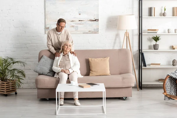 Senior husband standing near sick wife sitting on sofa at home — Stock Photo