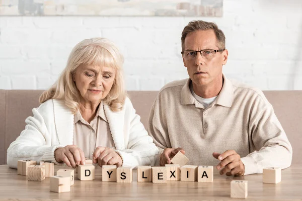 Засмучена жінка на пенсії, дивлячись на дерев'яні кубики з альцгеймером, що тече біля хворого чоловіка в окулярах — стокове фото