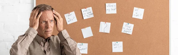 Panoramic shot of retired man with alzheimer disease touching head and standing near board with papers and letters — Stock Photo