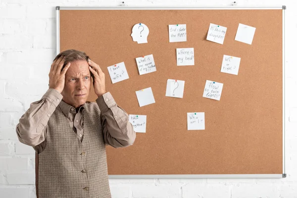 Retired man with alzheimer disease touching head and standing near board with papers and letters — Stock Photo