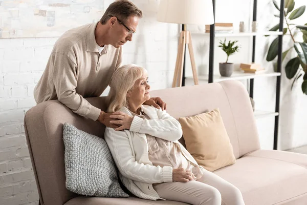 Trauriger älterer Mann steht neben kranker Frau und sitzt zu Hause auf Sofa — Stockfoto