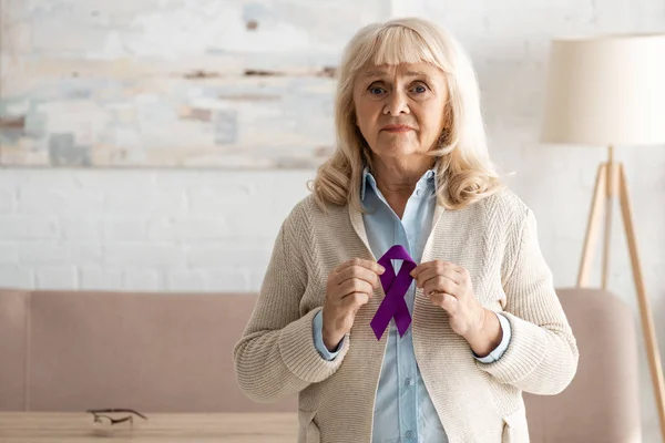 Frustrated and retired woman holding purple ribbon at home — Stock Photo