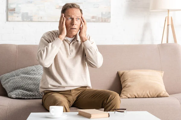 Jubilado con enfermedad mental tocando la cabeza mientras está sentado en el sofá — Stock Photo
