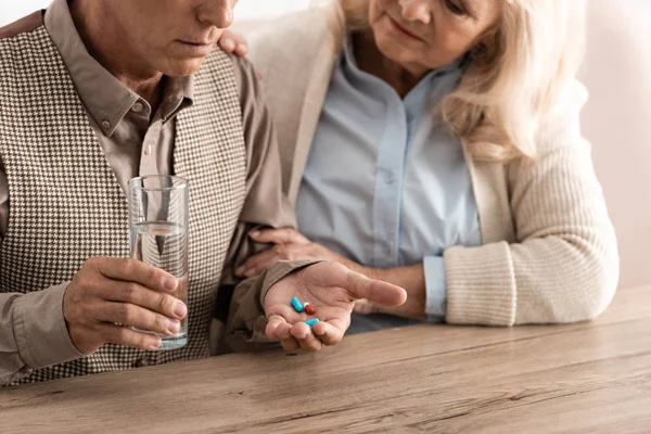 Visão cortada da mulher idosa sentada com o marido doente segurando vidro com água e pílulas — Fotografia de Stock