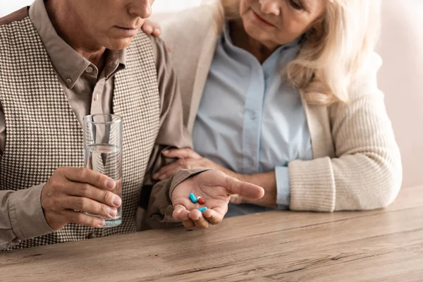 Vista recortada de la mujer mayor sentada con el marido enfermo sosteniendo vidrio con agua y pastillas - foto de stock