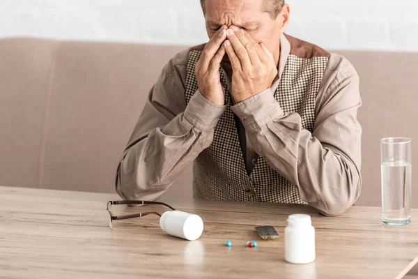 Hombre retirado molesto cubriendo la cara mientras está sentado cerca de píldoras en la mesa - foto de stock