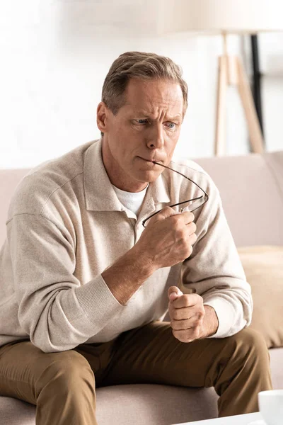 Thoughtful retired man with memory loss sitting on sofa and holding glasses — Stock Photo