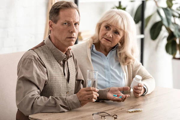 Senior femme assise avec mari malade tenant verre avec de l'eau et des pilules — Photo de stock