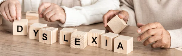 Panoramic shot of senior woman sitting near sick husband and wooden cubes with dyslexia letters — Stock Photo