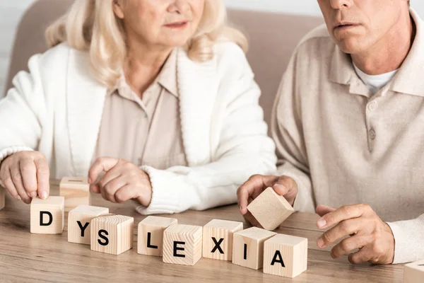 Vista recortada de la mujer mayor sentada cerca de marido enfermo y cubos de madera con letras de dislexia - foto de stock