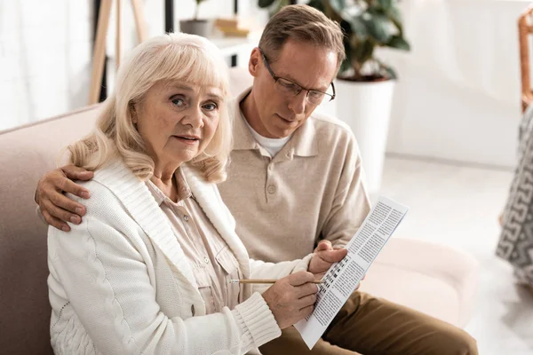 Senior man sitting near sick wife with alzheimer holding paper with letters and pencil — Stock Photo