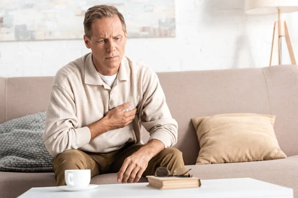 Sick retired man touching chest while sitting on sofa — Stock Photo