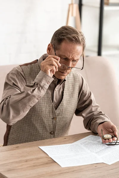 Senior man touching glasses while looking at papers and holding pocket watch — Stock Photo