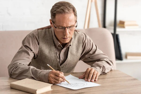 Uomo anziano con malattia mentale disegno su carta vicino libro a casa — Foto stock