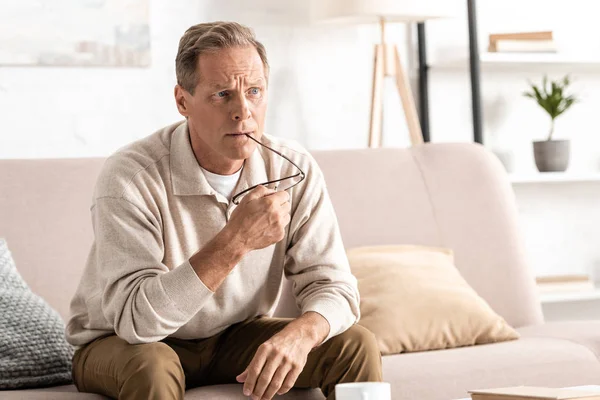 Pensive senior man with memory loss holding glasses and sitting on sofa — Stock Photo