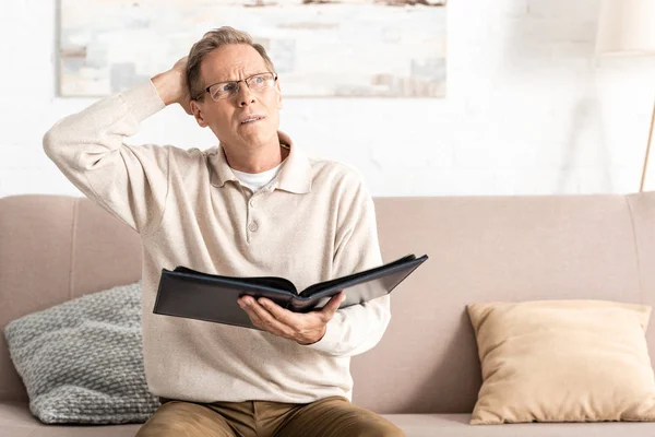 Confused senior man in glasses holding photo album while sitting on sofa — Stock Photo