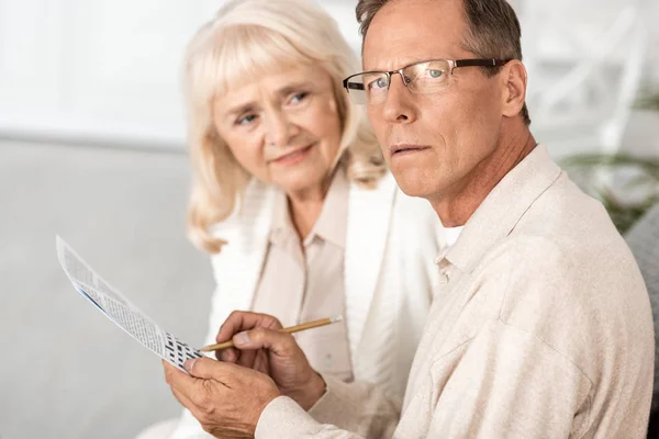 Селективное внимание больного пожилого человека с Альцгеймером, держащего карандаш рядом с кроссвордом и женой — стоковое фото