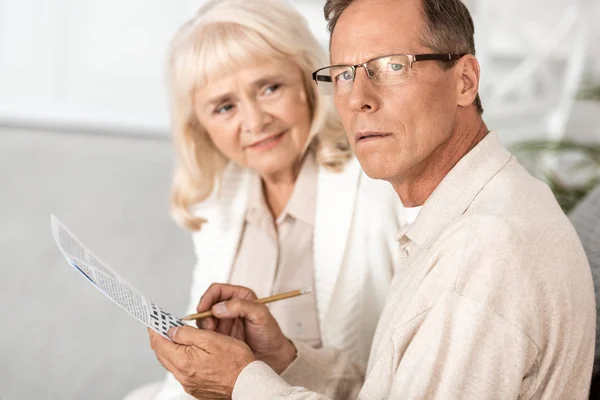 Selective focus of sick senior man with alzheimer holding pencil near crossword and wife — Stock Photo