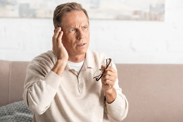 Thoughtful senior man with memory loss touching temple while holding glasses — Stock Photo