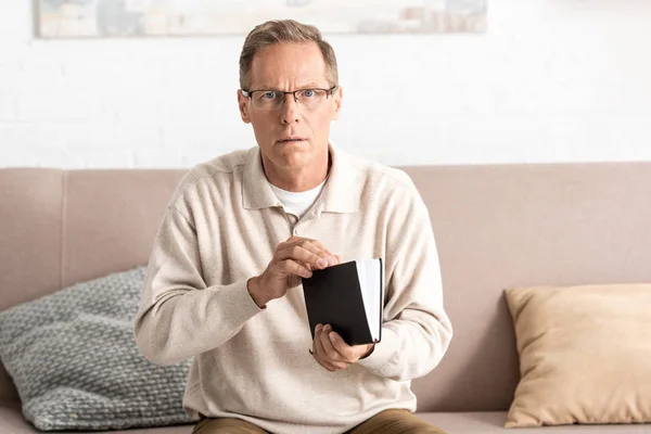 Senior man in glasses holding notebook while sitting on sofa — Stock Photo